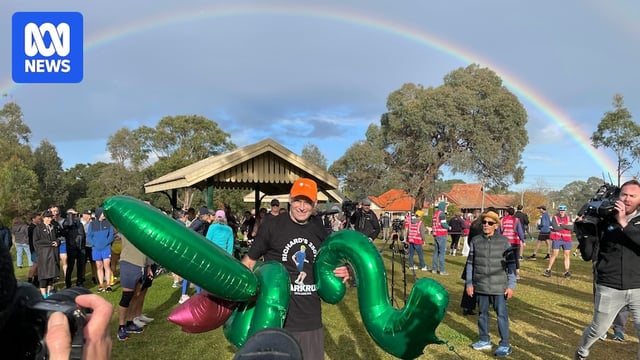 A man wearing an orange cap and black t-shirt stands in a park while holding green balloons with a rainbow in the background.
