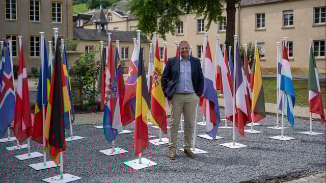 Michel Gloden, Bürgermeister von Schengen (Luxemburg), steht zwischen den Fahnen der Europäischen Union.