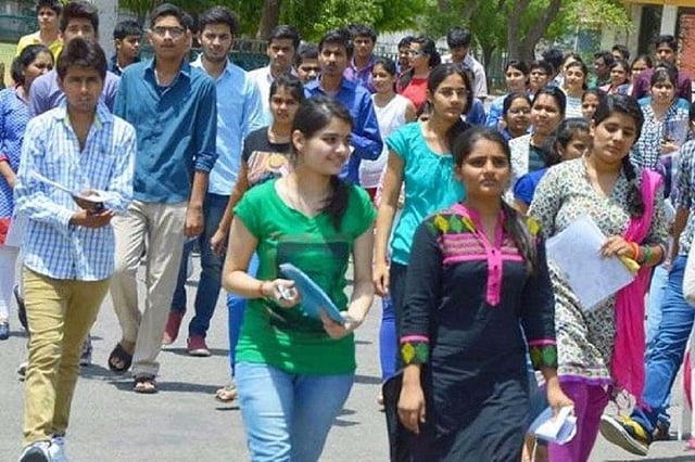 A large group of college-aged students, both men and women, are walking together outdoors in bright daylight. Most are casually dressed, holding papers or folders, suggesting they may be coming from or going to an exam or class. The atmosphere appears busy and energetic, with trees and a building visible in the background.