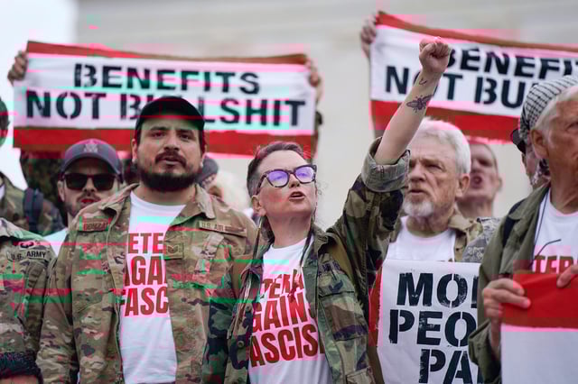 Protesters chant in front of a partially-obscured banner reading "BENEFITS NOT BULLSHIT" during an outdoor rally