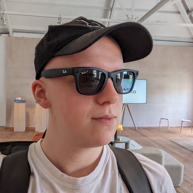 Hamish wearing a black pair of Wayfarer smart glasses from Ray-Ban and Meta. He's also wearing a hat and a bag in a large modern living room.