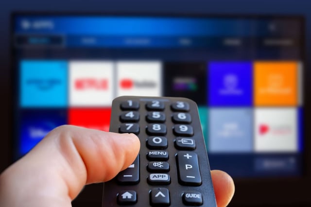 A man is holding a remote control of a smart TV in his hand. In the background, you can see the television screen with streaming entertainment apps for video on demand.