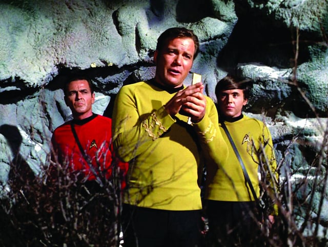 William Shatner as Captain James T. Kirk speaks into his communicator with James Doohan as Montgomery “Scotty” Scott and Walter Koenig as Pavel Chekov in the background during the episode “Spock’s Brain.”