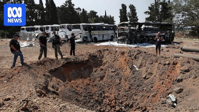 A large crater in dirt with burnt out buses behind.