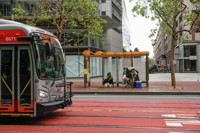 People take shelter at a bus stop as a Muni bus passes by on Market Street in San Francisco. Starting Saturday, multiple Muni lines will face service cuts.
