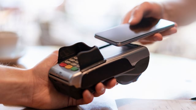 Eine Nahaufnahme einer kontaktlosen Bezahlvorgang. Eine Hand hält ein Bezahlterminal mit farbigen Tasten und sichtbarem Kartenschlitz, eine andere Hand hält ein Smartphone nahe an das Terminal.