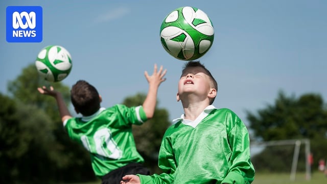 Two children in green are heading soccer balls in front of trees and blue sky.