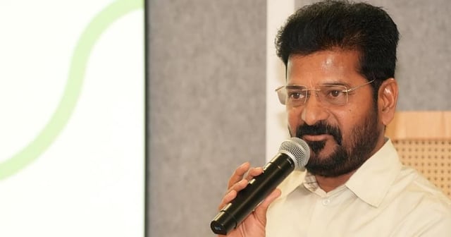Telangana CM Revanth Reddy in a cream shirt is speaking into a microphone while standing at a podium. He has short black hair, a beard, and is wearing rimless glasses. A laptop is placed on the podium in front of him. The background features a grey panel and a projection screen with a green curved line.









