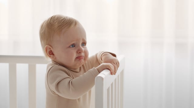 Little infant tired and hungry, start crying standing in crib, looking aside at empty space