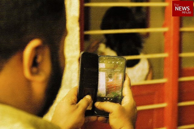 A man is seen secretly photographing a woman through a barred window using his smartphone. The woman, unaware and with her back turned, is indoors, while the man is outside focusing his phone camera on her. The image highlights an act of voyeurism or harassment, and includes a watermark from The News Minute in the top right corner.