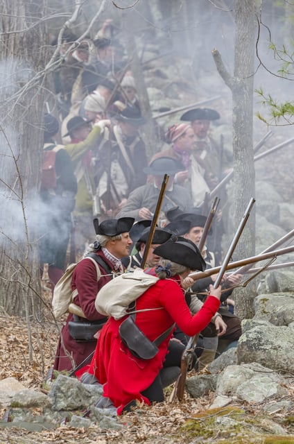 Revolutionary War reenactors in period dress crouch in a line with muskets behind a stone fence.
