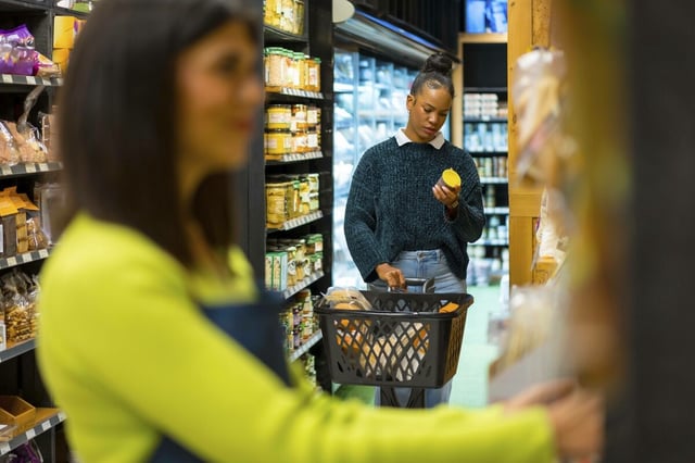 Eine Kundin wählt Bio Produkte aus, liest das Etikett und hält den Einkaufskorb im Supermarkt, während eine Verkäuferin die Regale ordnet