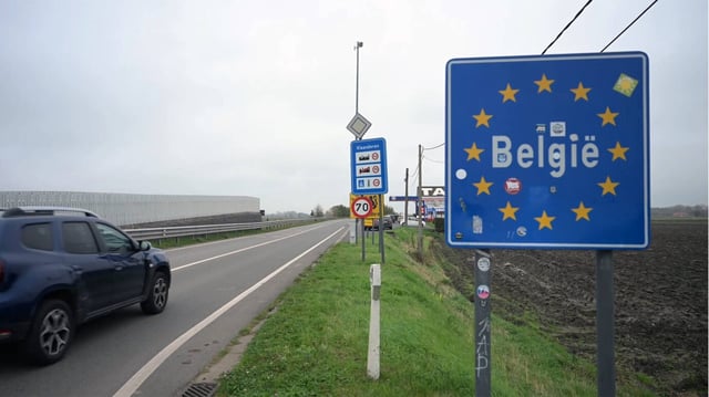 Straßenszene an der belgischen Grenze mit einem blauen Schild „België“ und zwölf gelben Sternen der EU, daneben weitere Verkehrsschilder, darunter ein Hinweis auf Flandern und ein Tempolimit. Ein Auto fährt in Richtung Landesinneres. Die Aufnahme entstand an einem bewölkten Tag und zeigt das flache Grenzgebiet mit Straße, Acker und Tankstelle. Grenzübertritt bei Flandern: Belgien will künftig wieder kontrollieren, wer ins Land fährt. Foto: IMAGO / Anadolu Agency