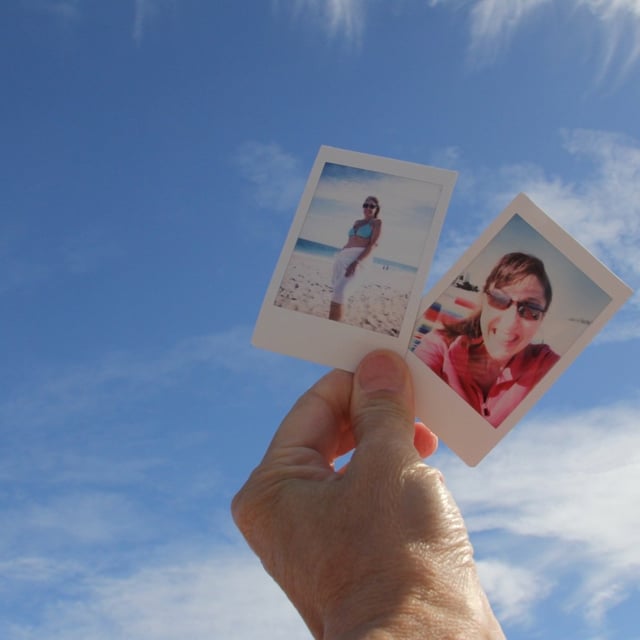 One hand holding up two instant photos of friends against blue sky