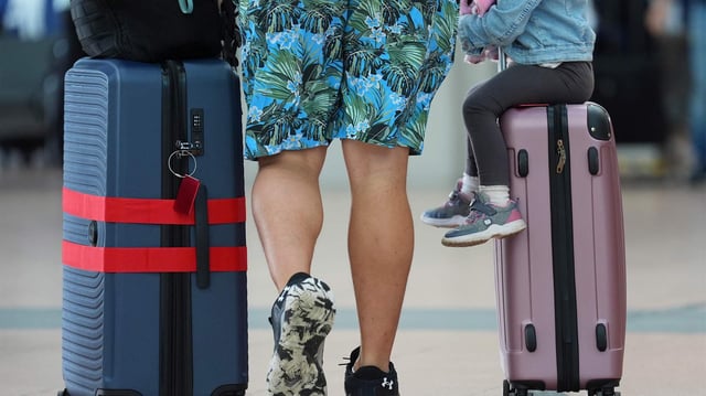 Ein Mann in Shorts mit Koffern am Flughafen. Auf einem Koffer sitzt ein Kind.