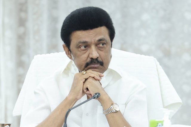 Tamil Nadu Chief Minister MK Stalin at a mic, dressed in a white shirt and seated before a white curtain.