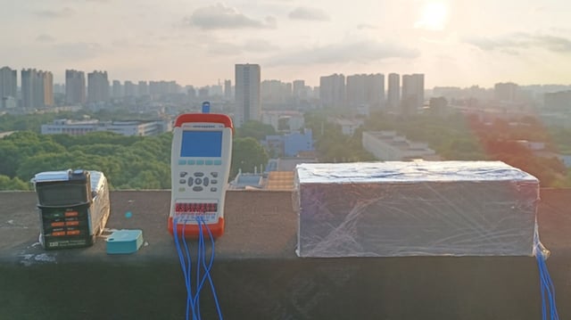 An outdoor experimental setup on a rooftop demonstrates radiative cooling performance. Instruments and sensors are connected to a block wrapped in bioplastic metafilm, with a dense city skyline and setting sun in the background—capturing real-world testing conditions for passive cooling technology.
