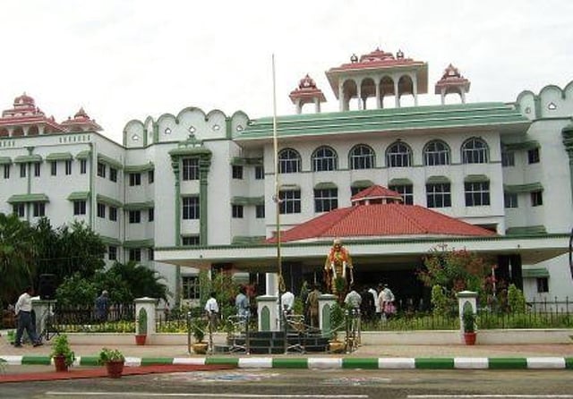 A file image of the Madurai Bench of the Madras High Court. The building is painted white and green. There is a red terracotta roof over the front portico. There is also a garlanded statue of MK Gandhi in front of the building. Several people can be seen walking about. There is a garden in front of the building. 