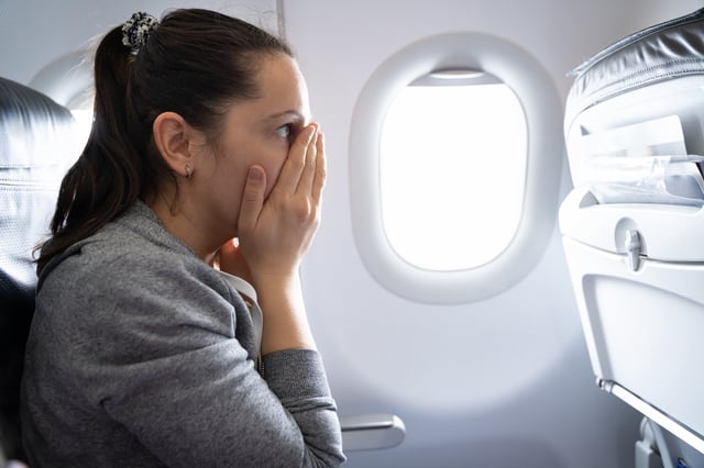 A stock image of a woman on a plane appearing nervous with her hands held over her face.
