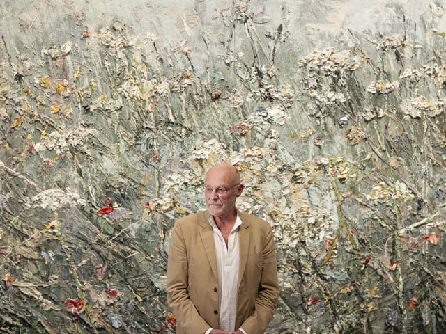 Artist Anselm Kiefer pictured in front of a textured, meadow-like painting