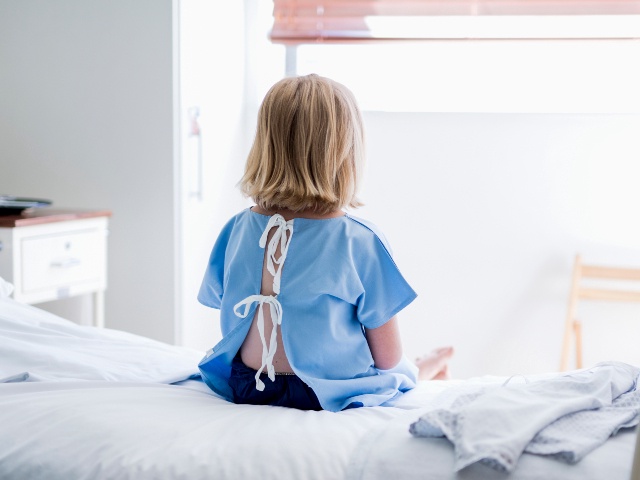 Rear view of ill girl sitting on bed. Female child patient is wearing hospital gown. She is resting in ward at clinic.