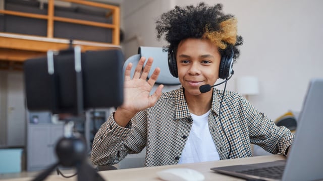 Portrait of teenage African-American boy wearing headset and waving at camera while streaming video games at home, young gamer or blogger concept, copy space