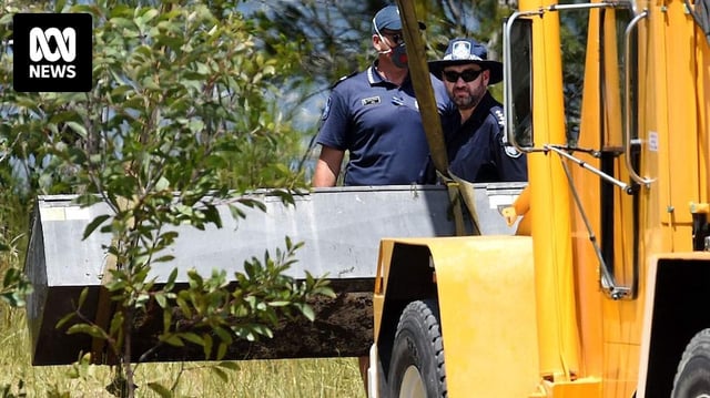 Two policemen watch as a yellow crane truck lifts a metal box in grassland.
