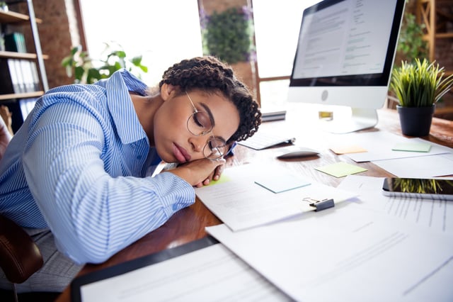 A stock image of an office worker taking a nap on her desk in front of a computer.