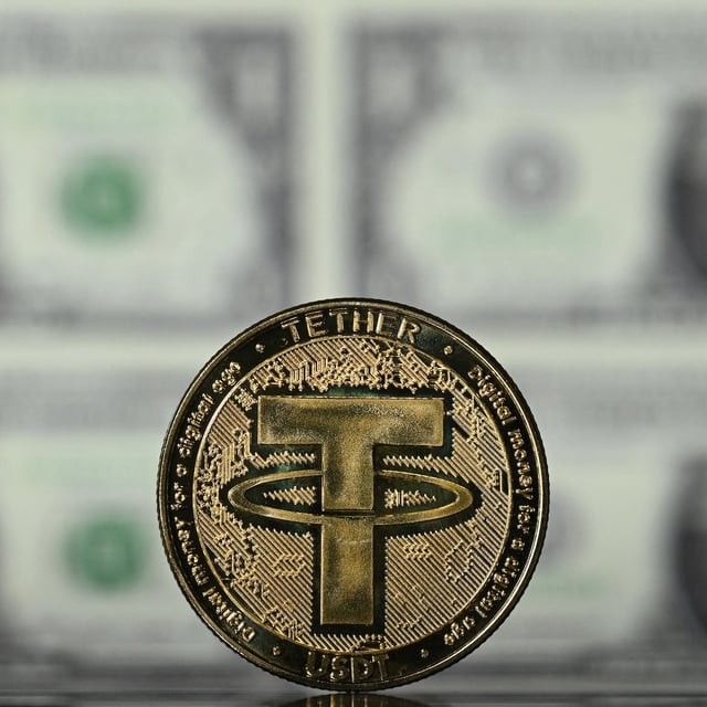 An illustration picture taken in London on May 8, 2022, shows a gold plated souvenir cryptocurrency Tether (USDT) coin arranged beside a screen displaying US dollar notes.