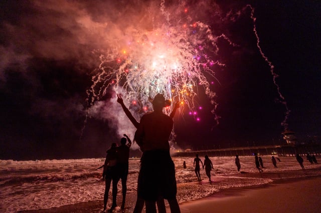 Huntington Beach, CA – July 04:  Couples Kellie Turner, of Huntington Beach and Peter Megariotis of Newport Beach, embrace at left, as Michael Marshall, of Paso Robles, and Carli Dugan, of San Louis Obispo, embrace at center, in the water as they watch the sky light up with fireworks over the ocean and pier on Independence Day, 4th of July in Huntington Beach Thursday, July 4, 2024.  (Allen J. Schaben / Los Angeles Times via Getty Images)