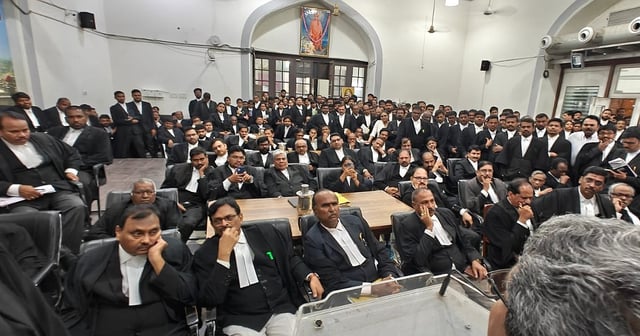 A large group of advocates, both men and women, dressed in black robes with white neckbands, are gathered in a courtroom or hall, seated and standing closely together. Most have serious or thoughtful expressions. The setting has arched doorways, a framed portrait on the wall, and air conditioning ducts visible above. A man with grey hair is seen from behind in the foreground, possibly addressing the group.