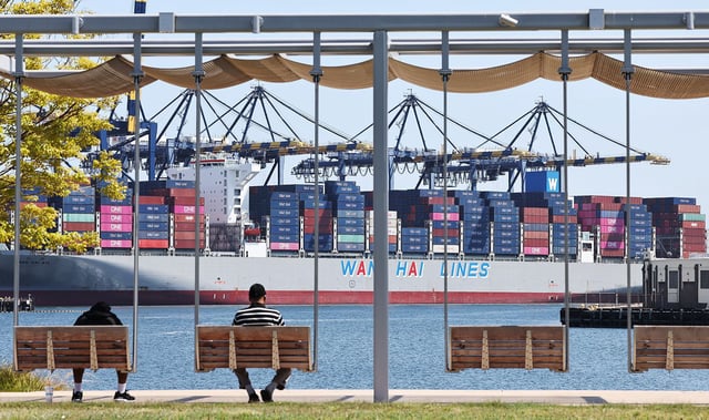People sit in a park near shipping containers stacked on a container ship at the Port of Los Angeles on June 25, 2025.