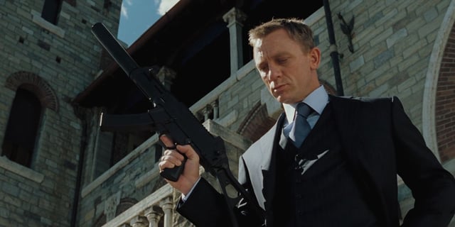 James Bond (Daniel Craig) stands before a villa wearing a sharp three-piece suit as he stands over a fallen adversary flaunting a cold glare and a sub-machine gun in 'Casino Royale' (2006).