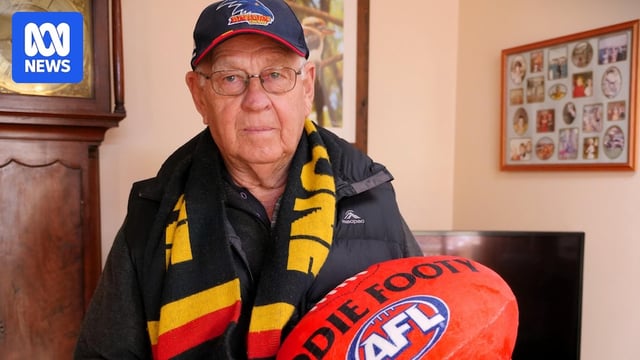 Peter Marks stands in front of his TV and two paintings, wearing an Adelaide Crows cap and scarf, holding a red toy football.