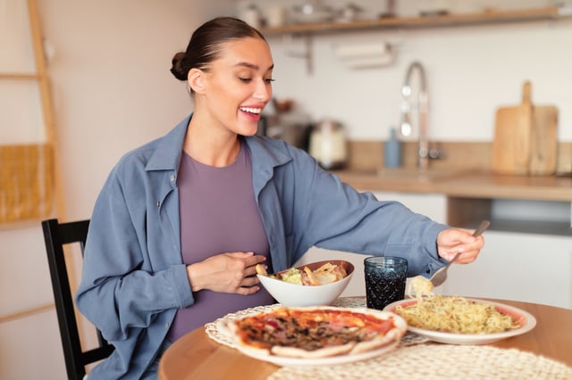 Hungry pregnant woman choosing from pizza, salad and eating pasta, having desire for junk food, sitting in kitchen.