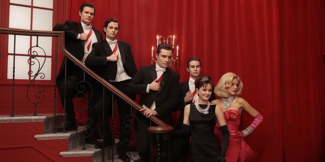Gossip Girl cast dressed in formal wear and posing for a picture on a staircase in front of a red curtain