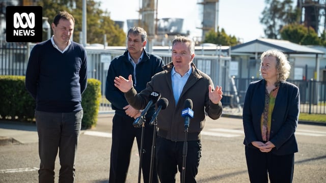 A middle-aged man gestures as he speaks near an industrial site.