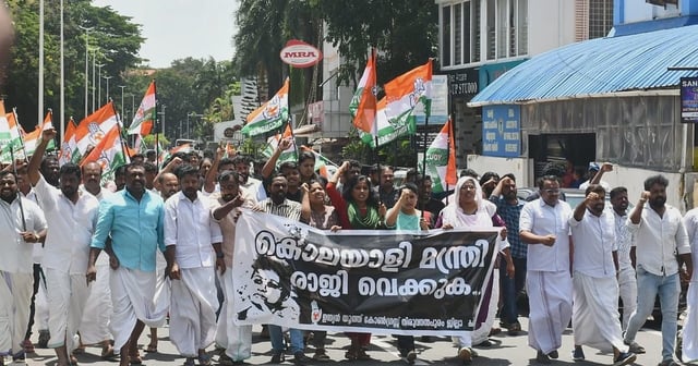 A large group of Youth Congress protestors march down a street in Thiruvananthapuram, holding a banner demanding the resignation of Health Minister Veena George. Many hold Congress flags and raise slogans, dressed in traditional Kerala attire.