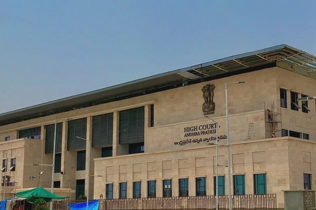 A wide view of the Andhra Pradesh High Court building, a modern structure with beige stone walls and large vertical black window shades. The Indian national emblem is mounted above the entrance, along with text in English and Telugu reading “HIGH COURT OF ANDHRA PRADESH.” Two workers are seen on scaffolding and a ladder on the side of the building, suggesting ongoing maintenance or construction work. A green canopy and some blue tarps are visible in the foreground.