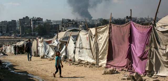 Des Palestiniens devant des tentes à Gaza-ville avec en arrière-plan une colonne de fumée s'élevant du centre de la bande de Gaza après un bombardement israélien, le 6 juillet 2025