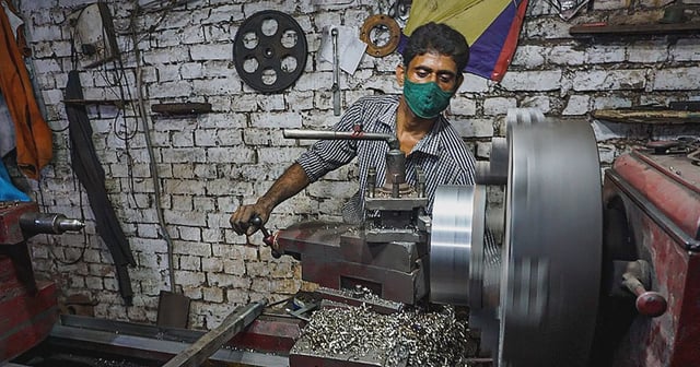 A man operates a lathe machine in a small workshop, wearing a striped shirt and a green face mask. Metal shavings are scattered around the machine, and the spinning metal component is in motion. The background features a white brick wall with hanging tools and machinery parts.