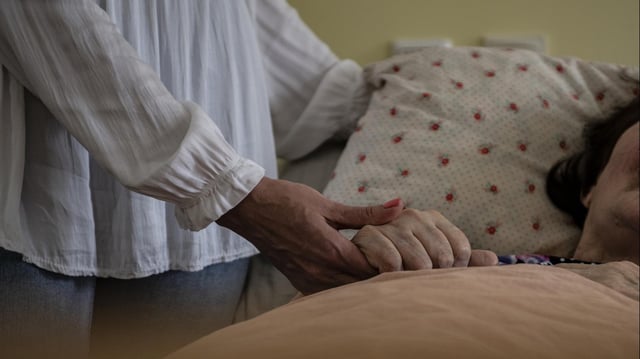  In einem Pflegeheim hält eine Frau die Hand einer Pflegebedürftigen Bewohnerin.