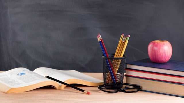 Desktop with books, pencils, apple, reading glasses and blank blackboard in background. Selective focus on front part of desk objects.