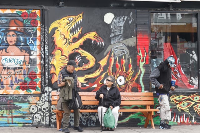 People stand next to and sit on a San Francisco Bay Area Bench Collective bench as they wait for a bus on 18th Street. The collective adds benches at or near bus stops without adequate seating.