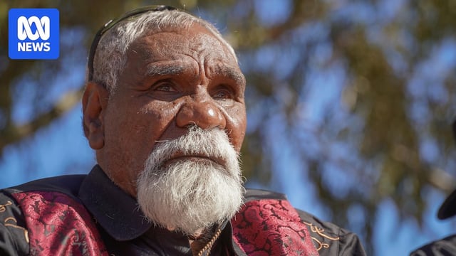 An Aboriginal man with a white moustache and beard, white hair, red vest over black shirt with a sombre expression.