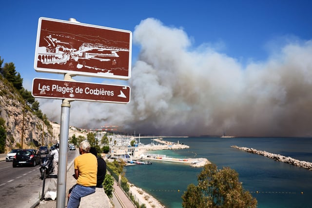 People look on near the Plage des Corbieres, on the outskirts of Marseille, southern France on July 8, 2025, as a smoke from a wildfire rages in the background.