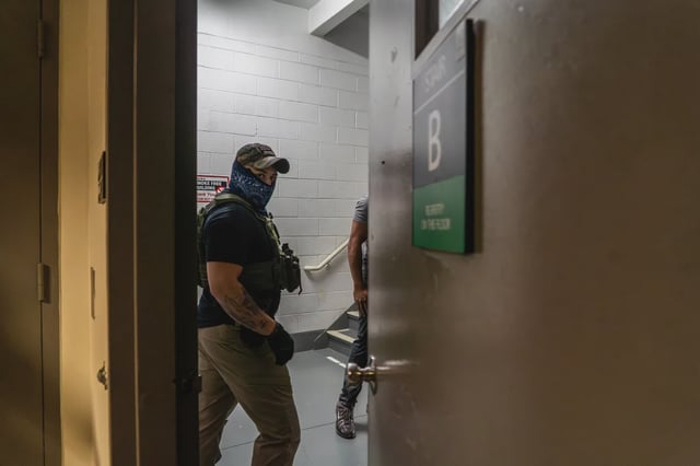 A masked federal agent looks directly at the camera as someone is detained and brought downstairs at 26 Federal Plaza.