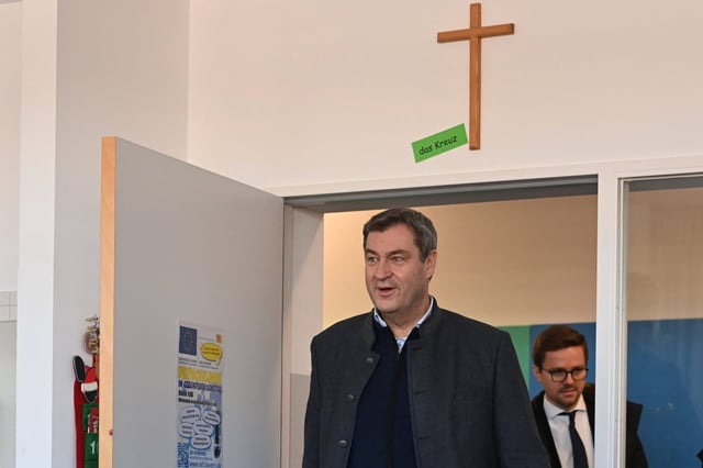 Bayerns Ministerpräsident Markus Söder steht in einem Schulgebäude unter einem an der Wand angebrachten Kreuz. Über dem Türrahmen hängt ein schlichtes Holzkruzifix mit dem Schild „das Kreuz“. Im Hintergrund ist ein Mitarbeiter zu sehen. Bayerns Ministerpräsident unter dem Kreuz: Ein Gericht urteilt gegen Kruzifixe in Schulen. Foto: IMAGO / Sven Simon