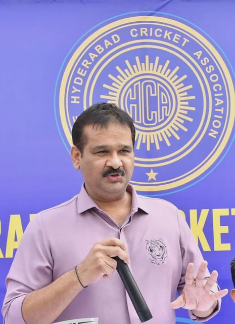 A man in a light purple shirt with a tiger emblem is speaking into a microphone at a public event. He stands in front of a blue and yellow backdrop featuring the logo of the Hyderabad Cricket Association (HCA), with "HCA" in the center and the full name encircling it.
