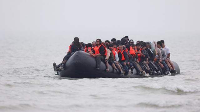 Eine Gruppe von Menschen, teils mit Rettungswesten, sitzt auf einem kleinen, schwarzen Schlauchboot.